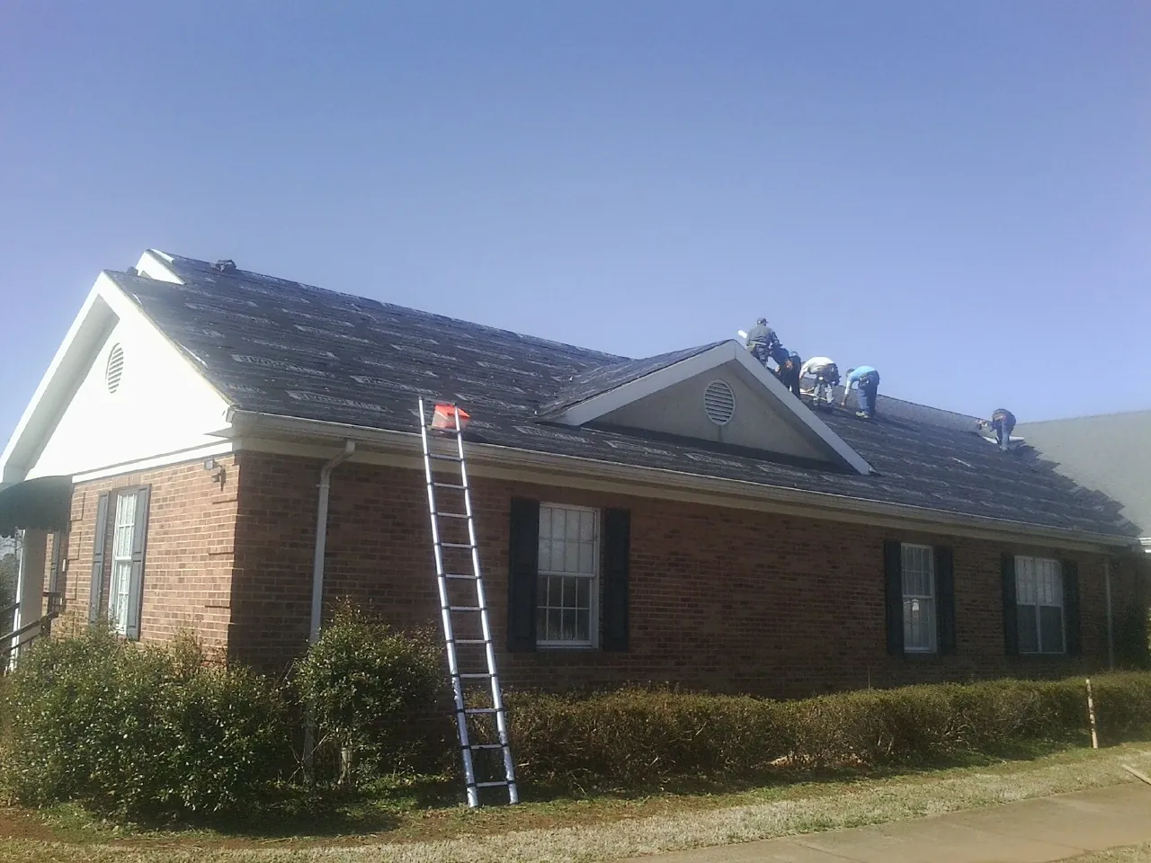 Crew tearing shingles off a brick ranch roof with a ladder set — Emergency Roof Repair work by our crew in South Barre