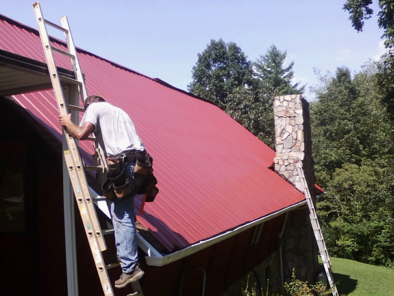 Roofer on a ladder beside a red metal roof and stone chimney — Soffit & Fascia Repair by Roofing Brothers in South Barre